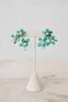 A pair of turquoise and green flower-shaped earrings with clear rhinestone centers, displayed on a cream fabric jewelry stand against a light neutral background.