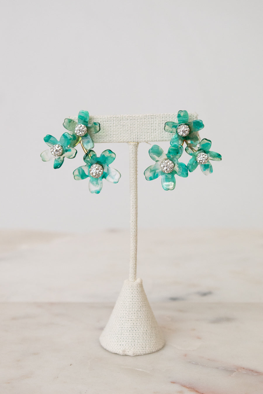 A pair of turquoise and green flower-shaped earrings with clear rhinestone centers, displayed on a cream fabric jewelry stand against a light neutral background.