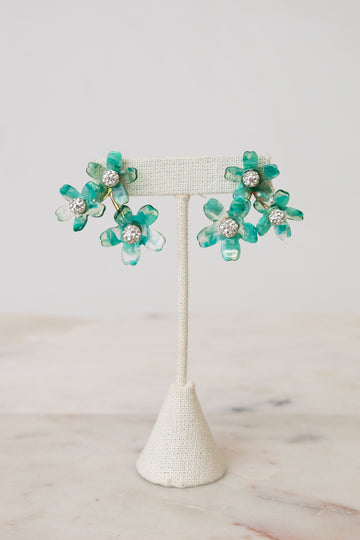 A pair of turquoise and green flower-shaped earrings with clear rhinestone centers, displayed on a cream fabric jewelry stand against a light neutral background.