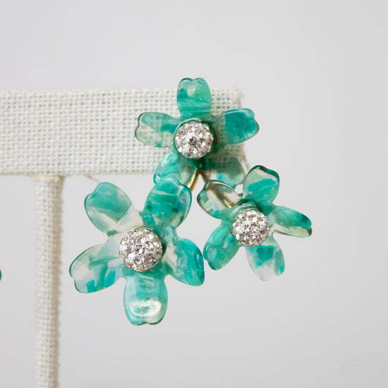 A pair of turquoise and green marbled flower-shaped earrings with clear rhinestone centers, displayed on a cream-colored jewelry stand against a light gray background.