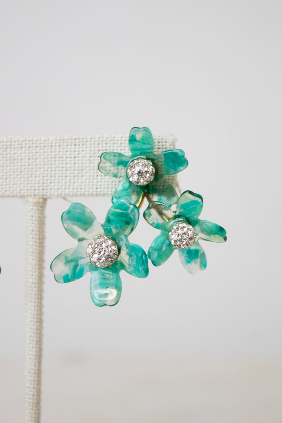 A pair of turquoise and green marbled flower-shaped earrings with clear rhinestone centers, displayed on a cream-colored jewelry stand against a light gray background.