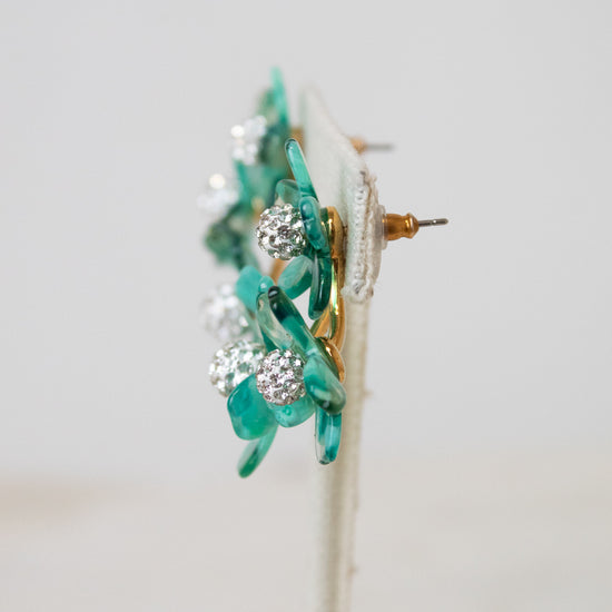 A pair of floral earrings with translucent green glass petals, clear rhinestone clusters, and gold-tone metal posts, displayed on a white stand against a light gray background.