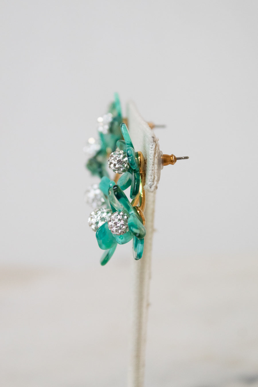 A pair of floral earrings with translucent green glass petals, clear rhinestone clusters, and gold-tone metal posts, displayed on a white stand against a light gray background.