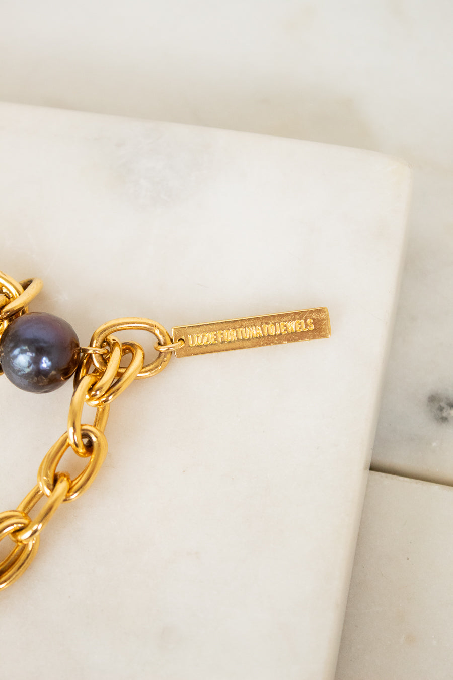 Gold chain bracelet with a pearl on a white surface, featuring the brand name 'Lizzie Fortunato'.