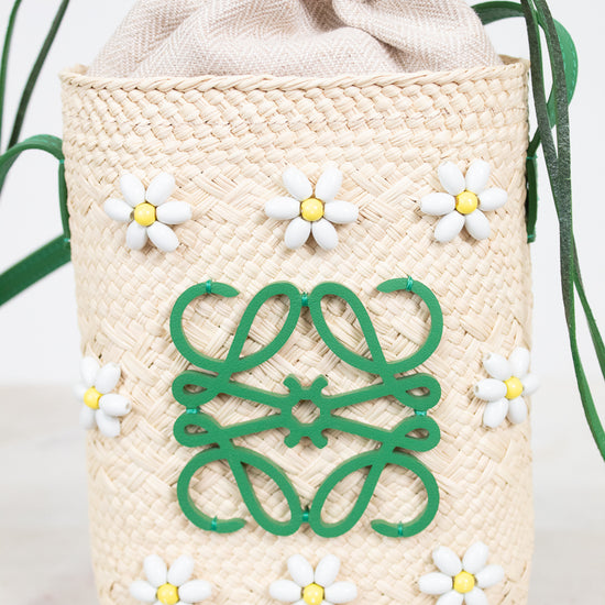 A woven straw drawstring bag in a warm cream color, decorated with a green Loewe anagram logo on the front, white daisy beaded embellishments, and green drawstring ties, set against a light marble background.