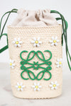 A woven straw drawstring bag in a warm cream color, decorated with a green Loewe anagram logo on the front, white daisy beaded embellishments, and green drawstring ties, set against a light marble background.