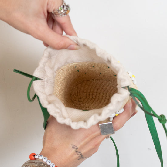 A close-up of a person's hands holding open a small woven straw bag with a cream fabric trim and green drawstring ties, against a plain white background.