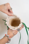 A close-up of a person's hands holding open a small woven straw bag with a cream fabric trim and green drawstring ties, against a plain white background.