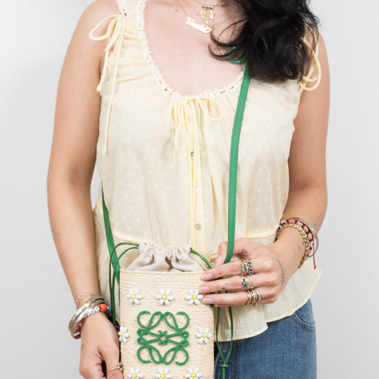 A woman wearing a cream-colored sleeveless blouse and blue jeans holds a small cream woven crossbody bag with a green logo and yellow and white flower accents. The background is a plain light gray studio wall.