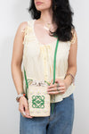 A woman wearing a cream-colored sleeveless blouse and blue jeans holds a small cream woven crossbody bag with a green logo and yellow and white flower accents. The background is a plain light gray studio wall.