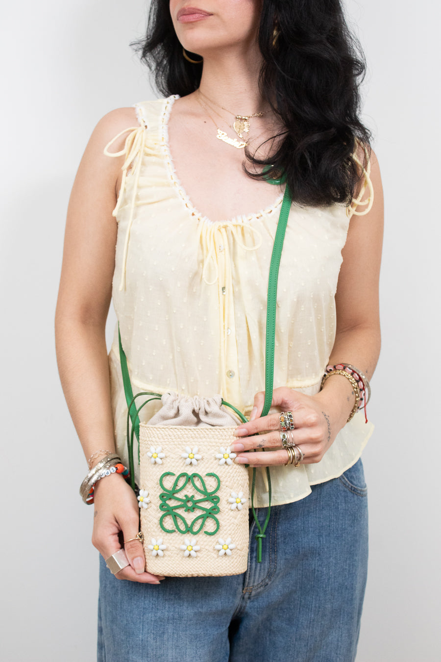 A woman wearing a cream-colored sleeveless blouse and blue jeans holds a small cream woven crossbody bag with a green logo and yellow and white flower accents. The background is a plain light gray studio wall.