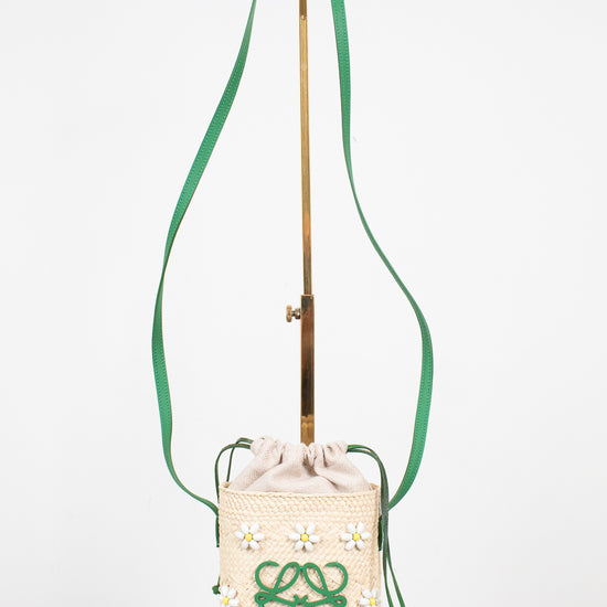 A cream woven bucket bag with a green logo and daisy pattern, featuring a green drawstring and a long green shoulder strap, displayed on a stand.