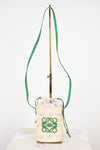A cream woven bucket bag with a green logo and daisy pattern, featuring a green drawstring and a long green shoulder strap, displayed on a stand.