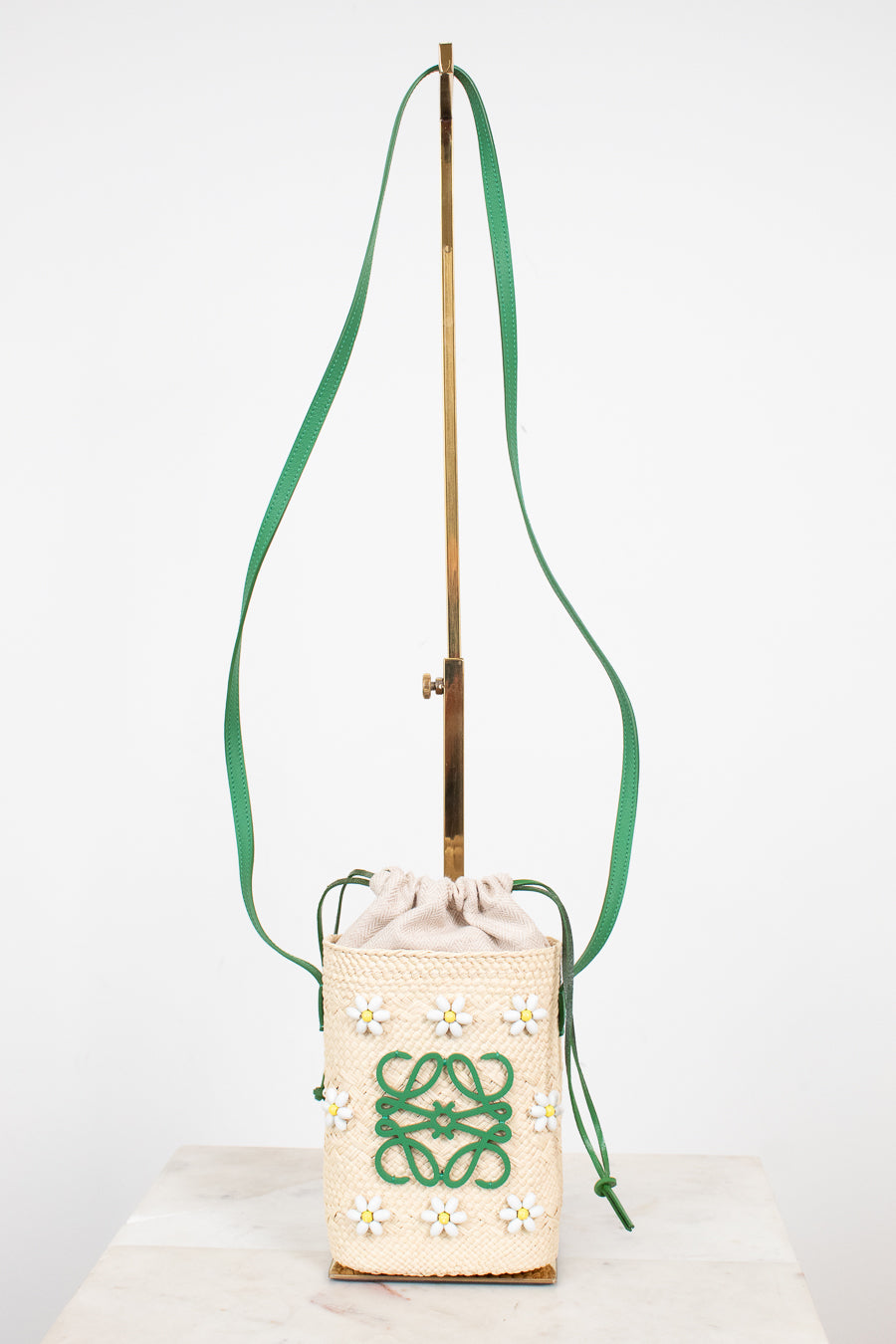 A cream woven bucket bag with a green logo and daisy pattern, featuring a green drawstring and a long green shoulder strap, displayed on a stand.