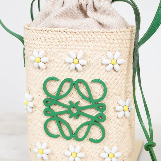 A woven straw bucket bag with a beige fabric and drawstring top, decorated with white and yellow daisy beaded appliqués and a green logo motif on the front. The bag is shown on a marble surface with a white background.