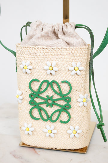 A woven straw bucket bag with a beige fabric and drawstring top, decorated with white and yellow daisy beaded appliqués and a green logo motif on the front. The bag is shown on a marble surface with a white background.