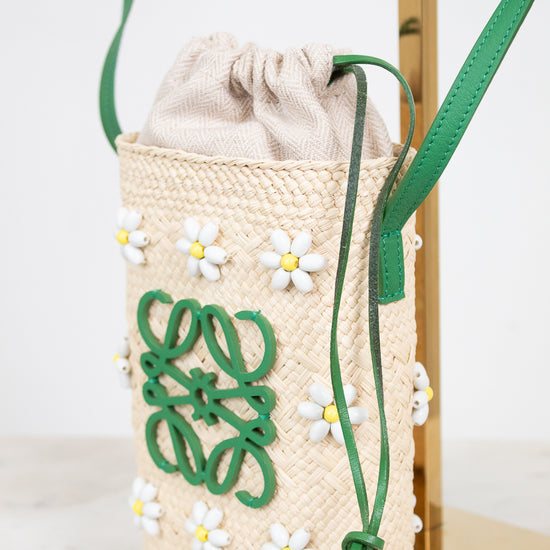 A woven straw drawstring bag with green leather strap and trim, decorated with white daisy appliqués and a green logo motif, displayed on a gold stand.