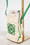 A woven straw drawstring bag with green leather strap and trim, decorated with white daisy appliqués and a green logo motif, displayed on a gold stand.