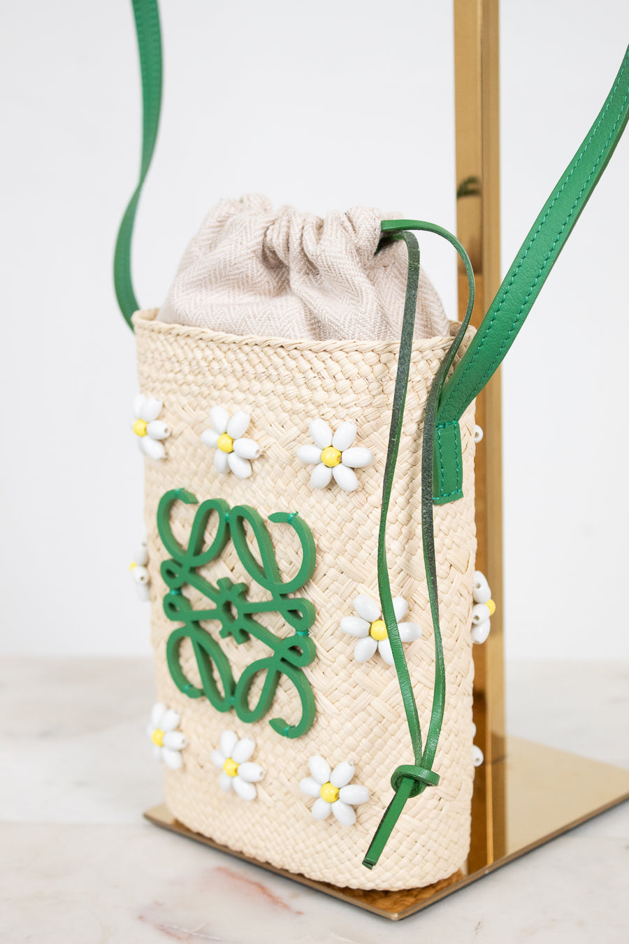 A woven straw drawstring bag with green leather strap and trim, decorated with white daisy appliqués and a green logo motif, displayed on a gold stand.