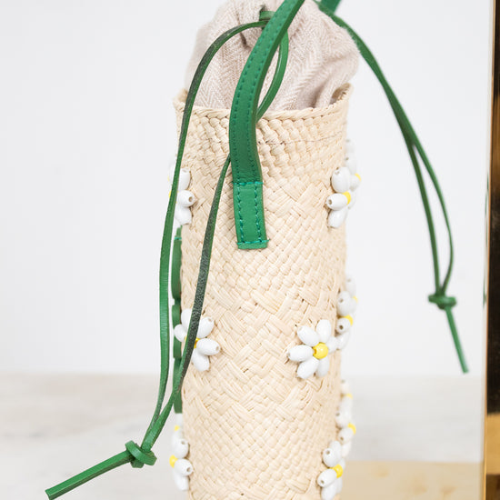 A woven straw bucket bag with green drawstring straps and white daisy beaded embellishments, displayed on a gold stand against a white background.