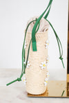 A woven straw bucket bag with green drawstring straps and white daisy beaded embellishments, displayed on a gold stand against a white background.