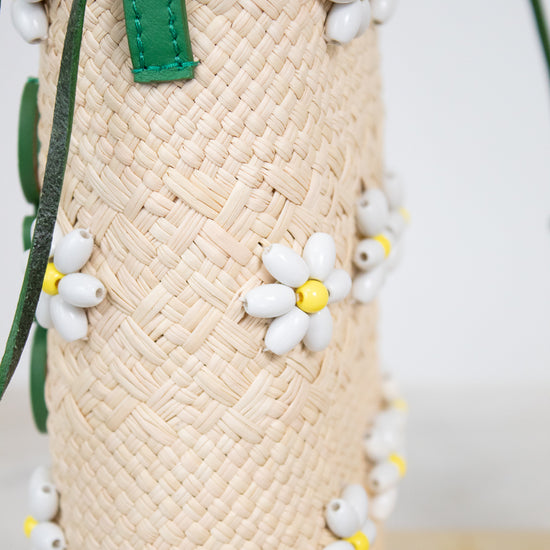 Close-up of a woven straw mini bucket bag with green leather strap accents and small white daisy beaded embellishments with yellow centers.