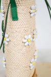 Close-up of a woven straw mini bucket bag with green leather strap accents and small white daisy beaded embellishments with yellow centers.