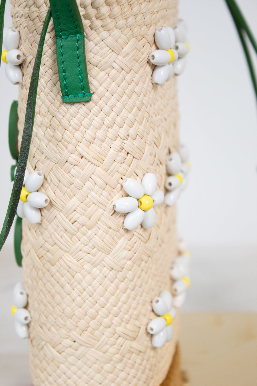 Close-up of a woven straw mini bucket bag with green leather strap accents and small white daisy beaded embellishments with yellow centers.