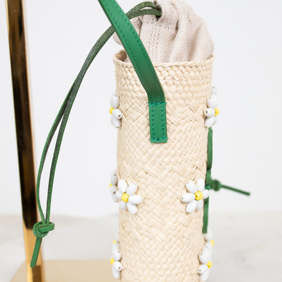A woven straw mini bucket shoulder bag in beige with green straps and small white daisy beaded  embellishments, displayed on a gold stand against a white background.