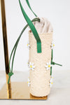A woven straw mini bucket shoulder bag in beige with green straps and small white daisy beaded  embellishments, displayed on a gold stand against a white background.