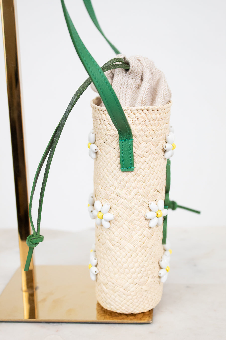 A woven straw mini bucket shoulder bag in beige with green straps and small white daisy beaded  embellishments, displayed on a gold stand against a white background.