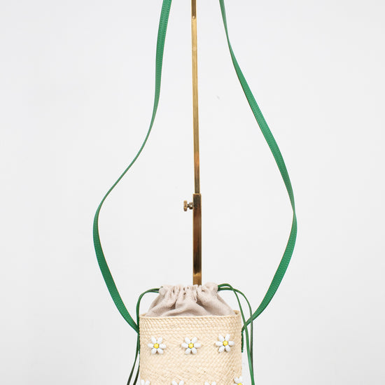 A cream woven bucket bag with embroidered daisy flowers, featuring a green shoulder strap and a gold-tone metal stand, set against a plain white background.