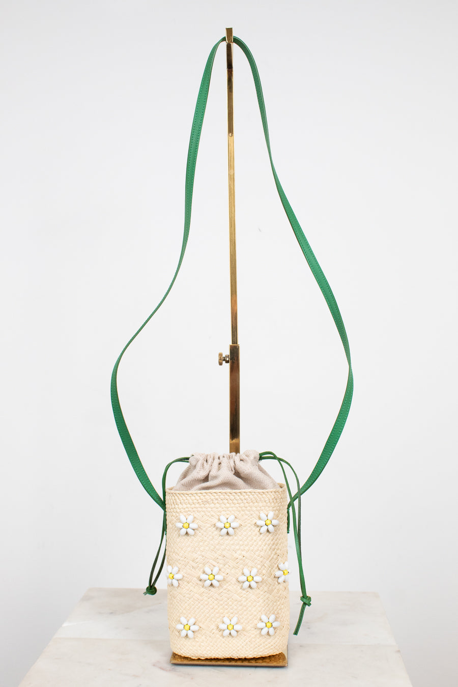 A cream woven bucket bag with embroidered daisy flowers, featuring a green shoulder strap and a gold-tone metal stand, set against a plain white background.