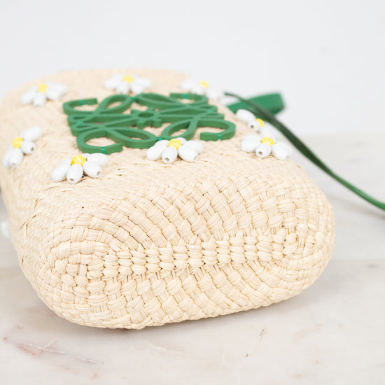 Bottom view of a woven straw bag in a natural beige color, decorated with a green logo detail and small white and yellow beaded  daisy flowers, resting on a light marble surface.