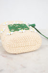 Bottom view of a woven straw bag in a natural beige color, decorated with a green logo detail and small white and yellow beaded  daisy flowers, resting on a light marble surface.