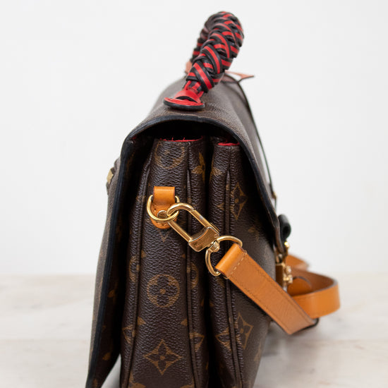 Brown monogrammed handbag with a red and black handle on a white background