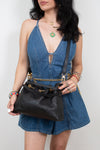 A woman wearing a blue denim romper and holding a black leather shoulder bag with gold chain accents, photographed against a plain light gray background.