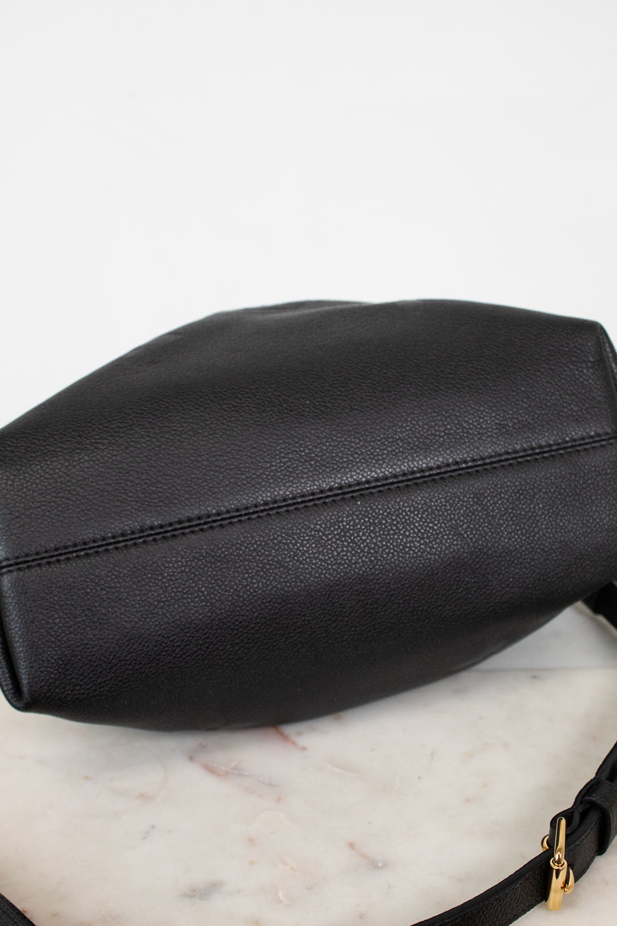 Bottom view of a black leather shoulder bag , resting on a white marble surface against a white background.