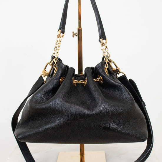A black leather bucket bag with gold-tone hardware, displayed on a gold stand against a white background.
