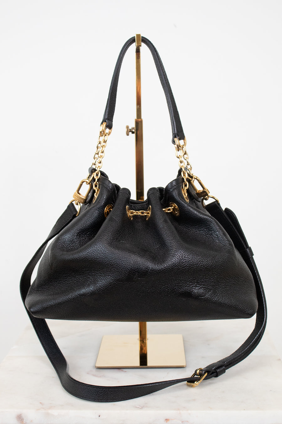 A black leather bucket bag with gold-tone hardware, displayed on a gold stand against a white background.