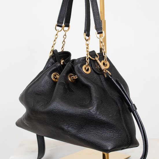 A black leather bucket bag with gold-tone hardware, displayed on a gold metal stand against a white background.