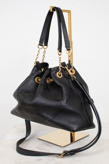 A black leather bucket bag with gold-tone hardware, displayed on a gold metal stand against a white background.