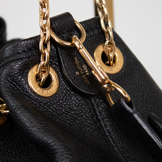 Close-up of a black leather handbag with gold-tone chain details and hardware, showing a small gold-tone logo plate and a black leather tag with gold lettering.