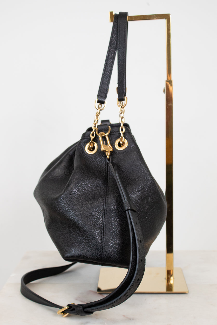 A black leather bucket bag with gold-tone hardware, displayed on a gold metal stand against a white background.