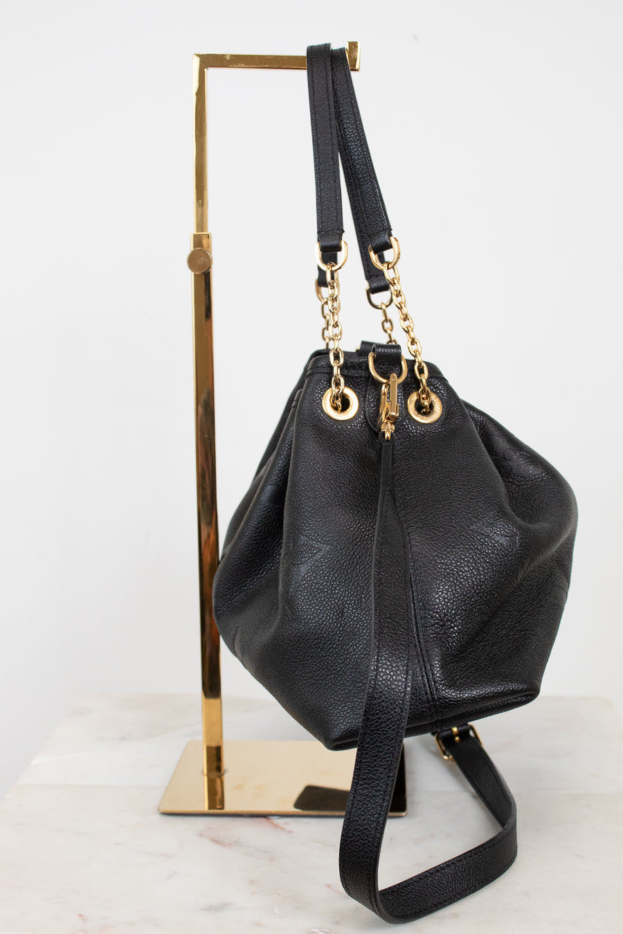 A black leather bucket bag with gold-tone hardware, displayed on a gold metal stand against a white background.