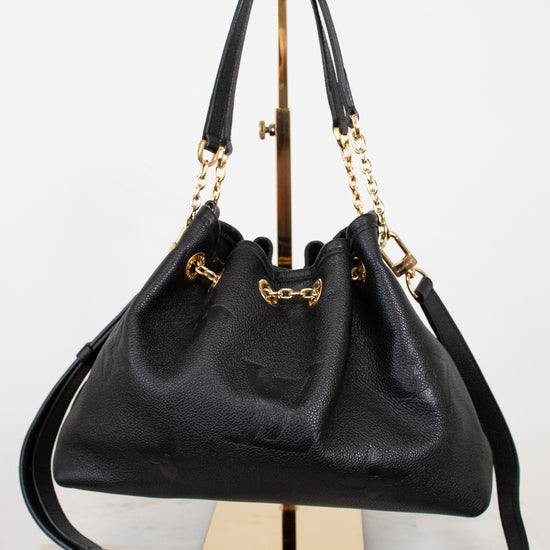 A black leather drawstring chain handbag with gold-tone hardware, displayed on a gold stand against a white background.