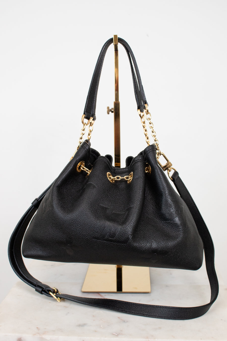 A black leather drawstring chain handbag with gold-tone hardware, displayed on a gold stand against a white background.
