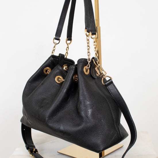 A black leather drawstring chain handbag with gold-tone hardware, displayed on a gold-tone metal stand against a white background.