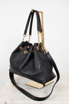 A black leather drawstring chain handbag with gold-tone hardware, displayed on a gold-tone metal stand against a white background.