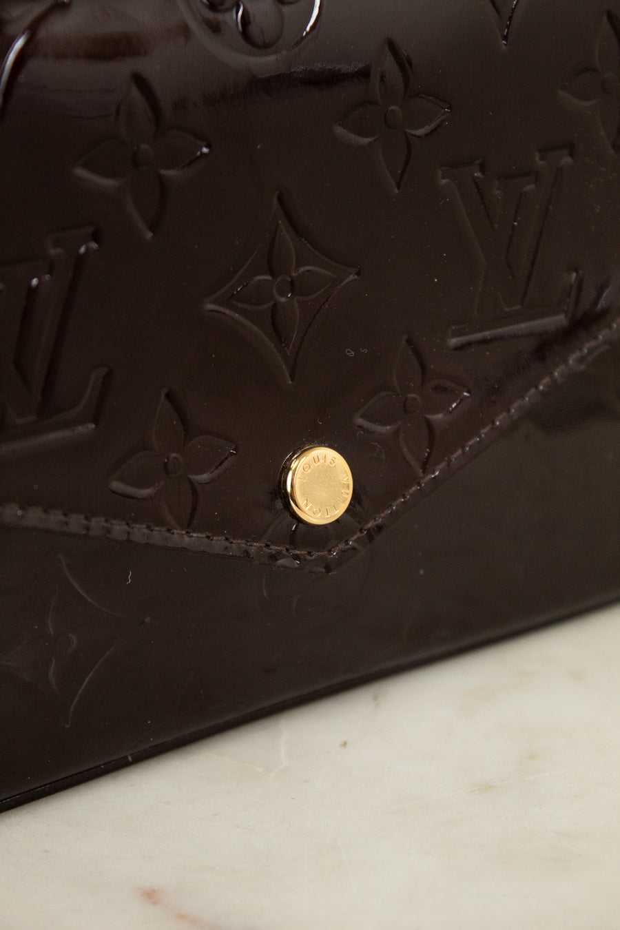 Close-up of a dark burgundy Louis Vuitton monogram leather bag with a gold-tone snap button, resting on a light marble surface.